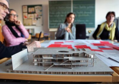 Im Vordergrund ein Pappmodell der ersten Architekturvorschläge, im Hintergrund sitzen Menschen an Tisch und blicken auf Modell.