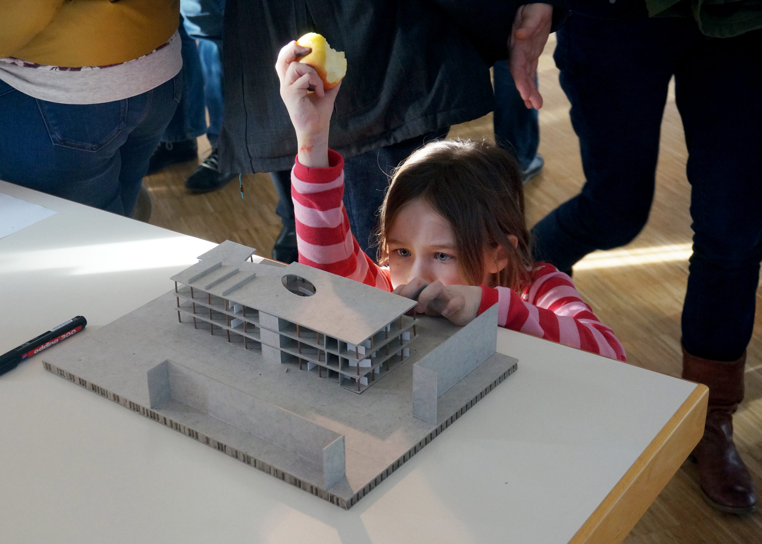 Junge Besucherin steht auf Augenhöhe mit Architekturmodell und betrachtet es. Dabei hält sie einen angebissenen Apfel in die Luft.